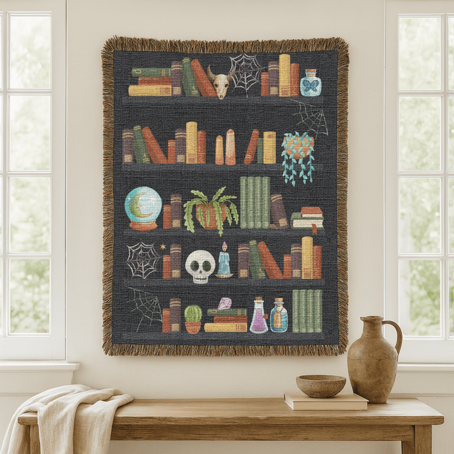 Charcoal fringed woven blanket printed as five shelves of books with a cow skull, cobwebs, a crystal ball, a trailing pumpkin, a candlelit skull and rows of potion bottles.