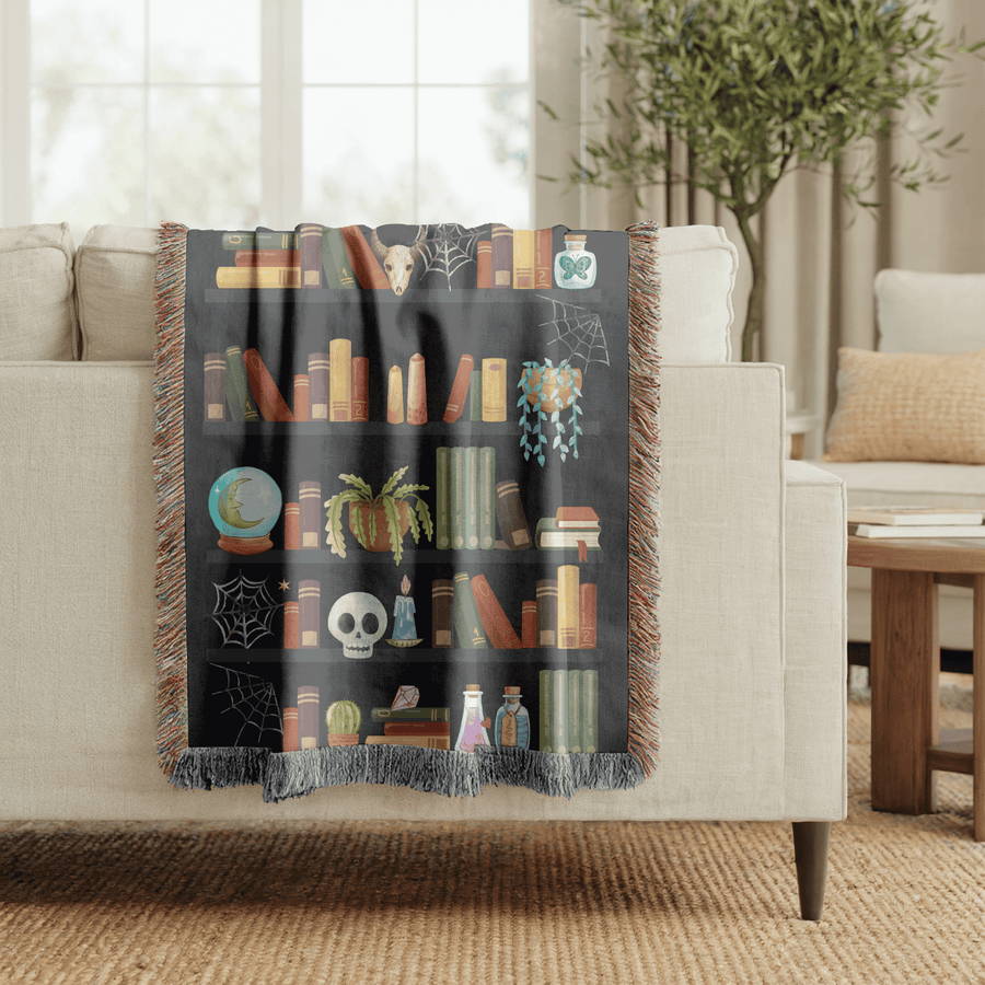 Charcoal fringed woven blanket printed as five shelves of books with a cow skull, cobwebs, a crystal ball, a trailing pumpkin, a candlelit skull and rows of potion bottles.
