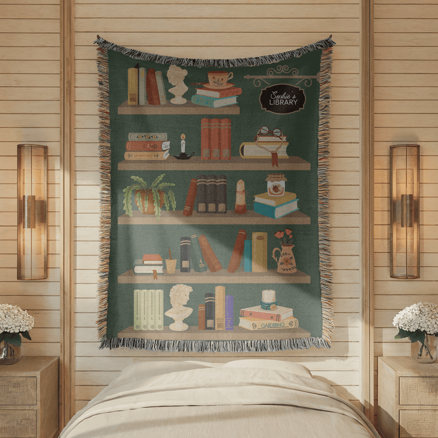 Teal fringed woven blanket printed as four bookshelves holding books, a teacup, a cat, a trailing plant, a candle and a vase of poppies, with a sign reading Sadie's Library.