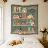 Teal fringed woven blanket printed as four bookshelves holding books, a teacup, a cat, a trailing plant, a candle and a vase of poppies, with a sign reading Sadie's Library.