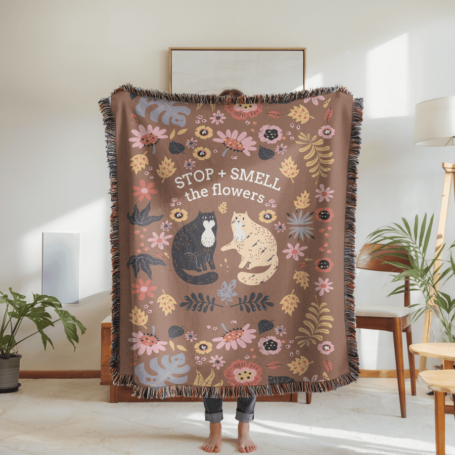 Brown fringed woven blanket reading Stop and Smell the Flowers above a black cat and a cream cat sitting among folk-art flowers, leaves and dark seed heads in pink, blue and gold.