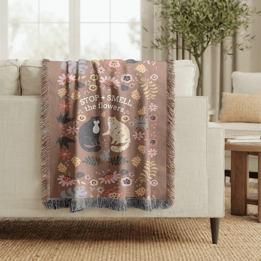 Brown fringed woven blanket reading Stop and Smell the Flowers above a black cat and a cream cat sitting among folk-art flowers, leaves and dark seed heads in pink, blue and gold.