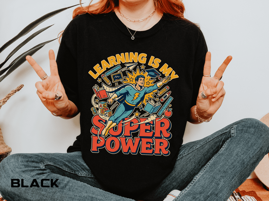 A black T-shirt printed with a comic-style teacher superhero flying among books and school supplies under Learning Is My Super Power.