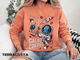 Terracotta sweatshirt printed with a blue alien in a spacesuit raising a coffee cup beside a rocket and a stovetop pot, headed Galactic Coffee, with cups and beans drifting around them.