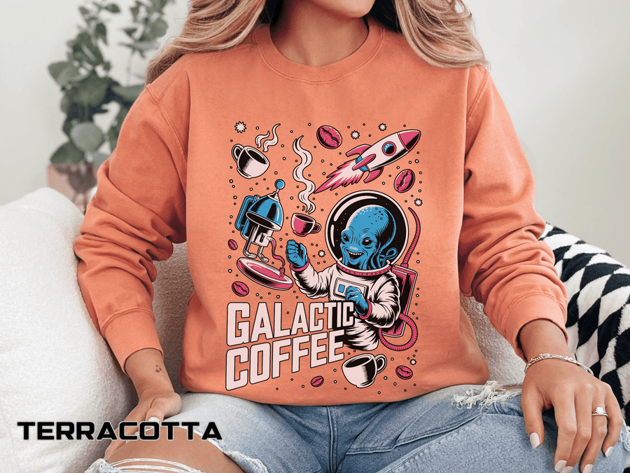 Terracotta sweatshirt printed with a blue alien in a spacesuit raising a coffee cup beside a rocket and a stovetop pot, headed Galactic Coffee, with cups and beans drifting around them.