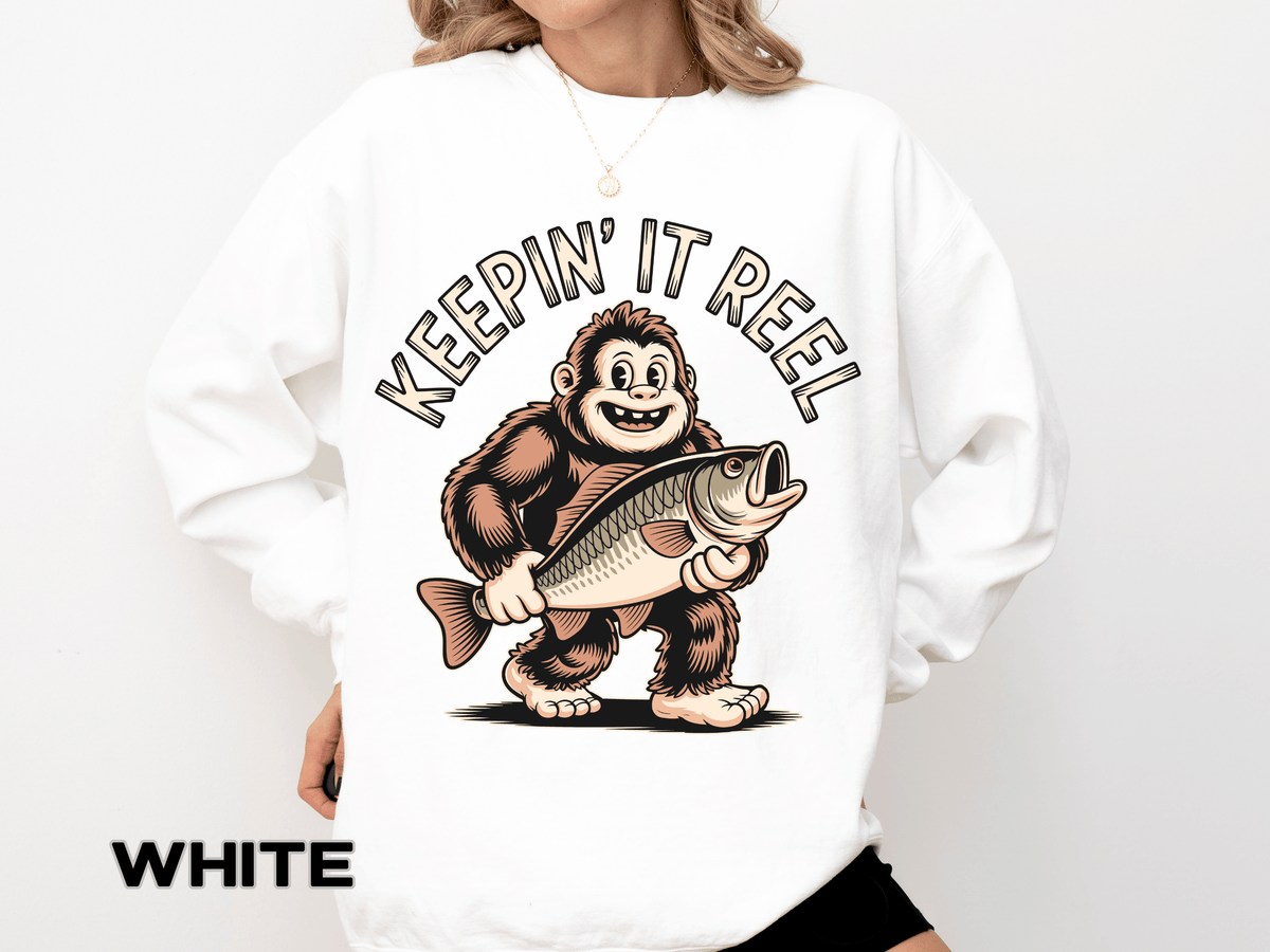 White sweatshirt printed with a grinning bigfoot carrying a large open-mouthed fish under one arm, headed Keepin It Reel in an arc of outlined capitals curving above his head.