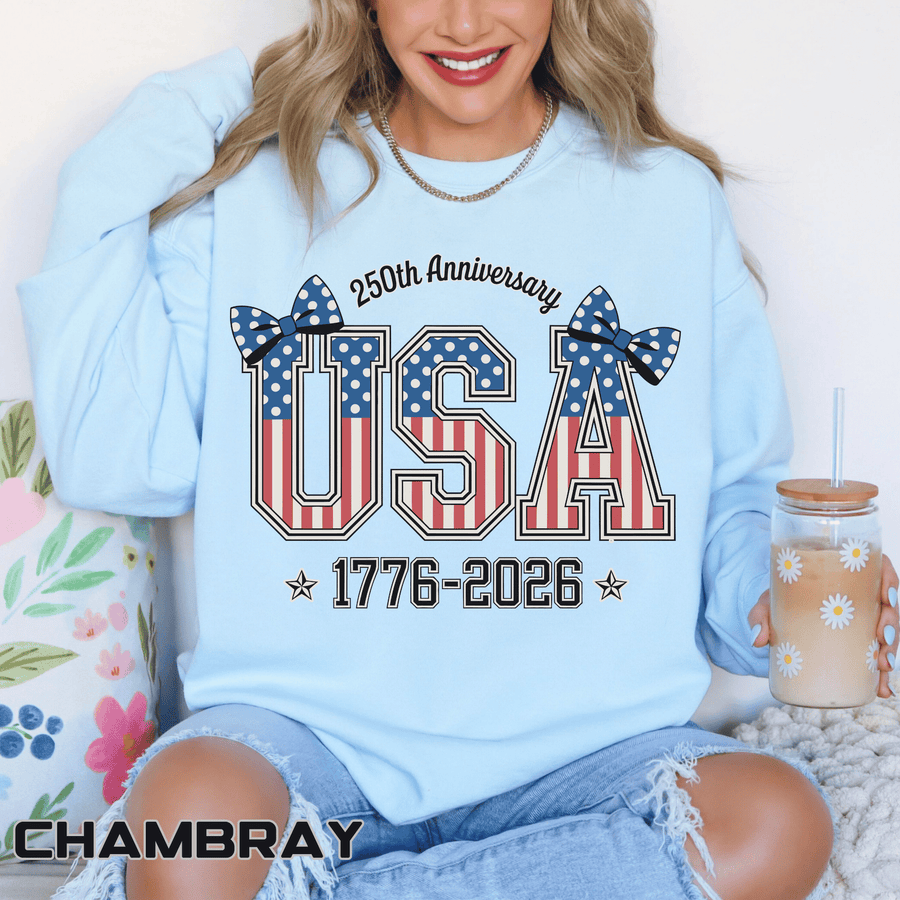 Chambray sweatshirt printed with USA in tall letters filled with the American flag, topped by two polka-dot bows, above the dates 1776 to 2026 and headed 250th Anniversary.