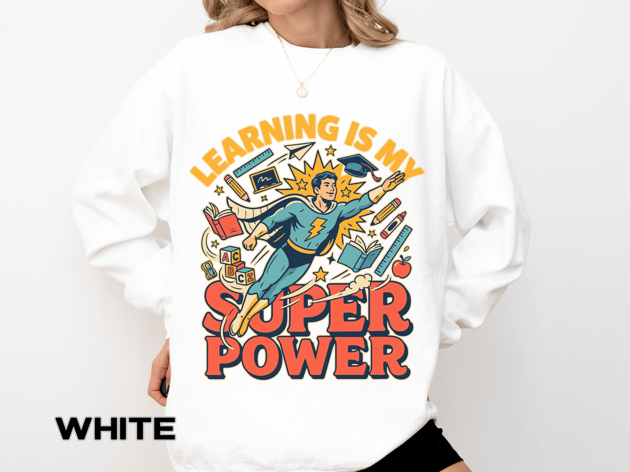 White sweatshirt printed with a comic-style teacher flying like a superhero in a cape and mortarboard through books and pencils, headed Learning Is My Super Power in orange.