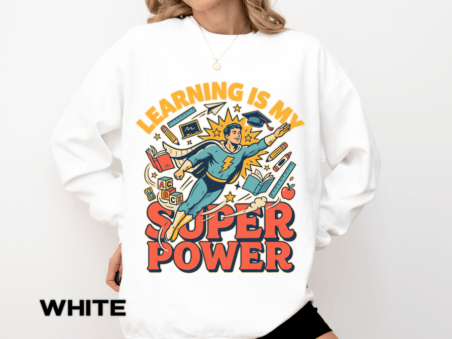 White sweatshirt printed with a comic-style teacher flying like a superhero in a cape and mortarboard through books and pencils, headed Learning Is My Super Power in orange.