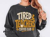 Pepper sweatshirt printed with the words Tired Teachers Coffee Club in chunky cream and gold lettering, a steaming mug set into the text and coffee beans scattered around.