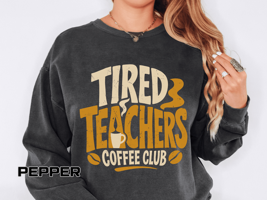 Pepper sweatshirt printed with the words Tired Teachers Coffee Club in chunky cream and gold lettering, a steaming mug set into the text and coffee beans scattered around.