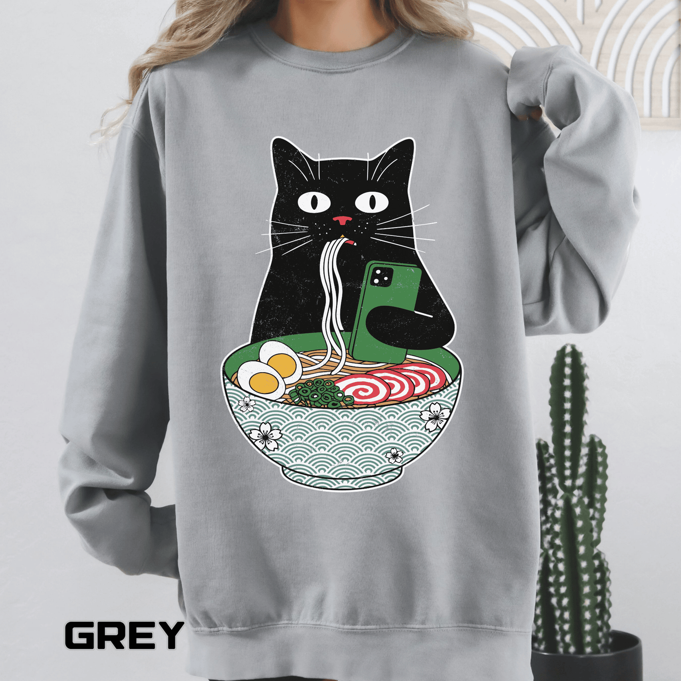 Grey sweatshirt printed with a black cat slurping noodles from a patterned ramen bowl while holding up a green phone, with egg, fish cake and spring onion in the broth below.