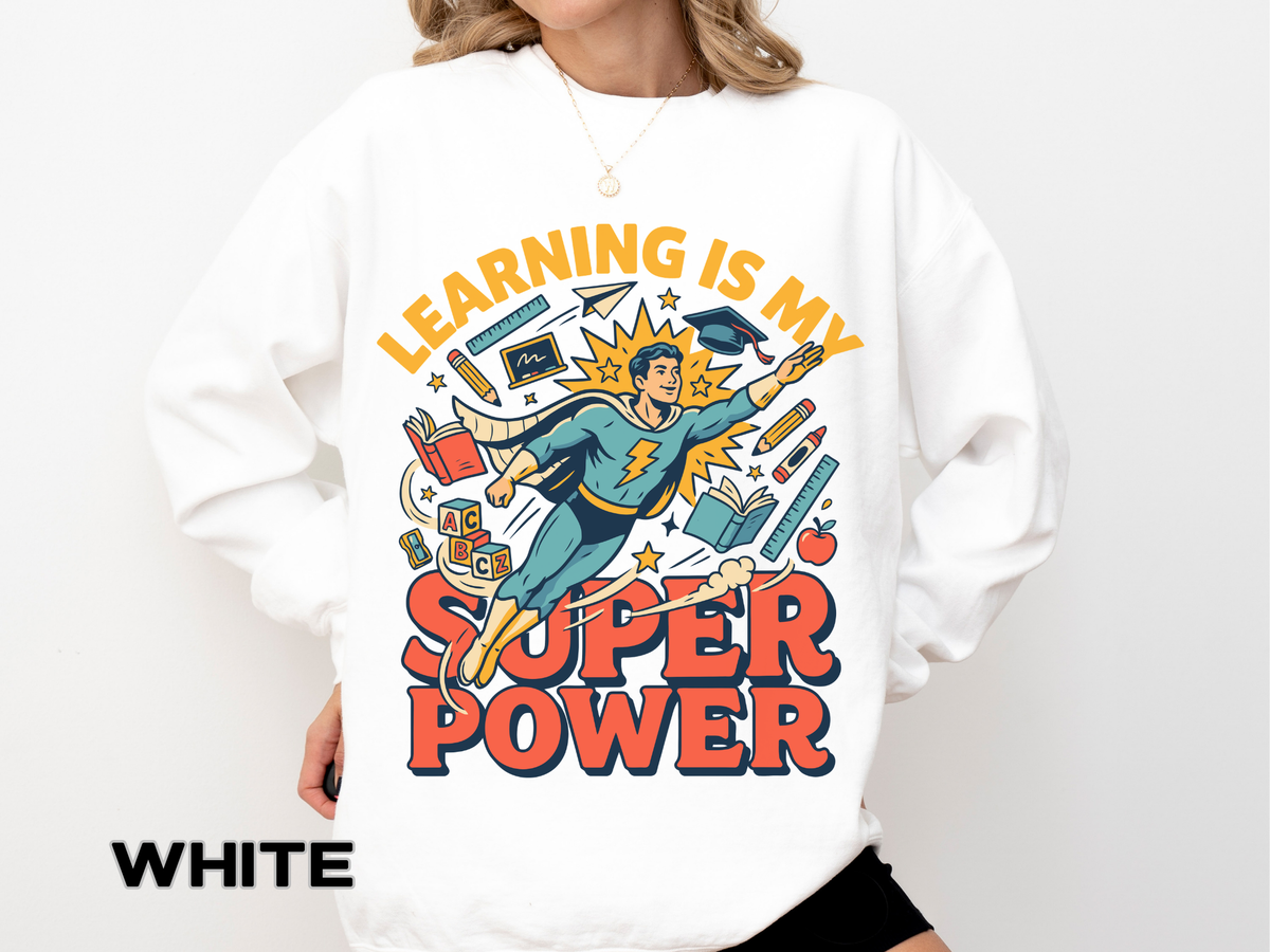 A white crewneck sweatshirt printed with a comic-style teacher superhero flying among books and school supplies under Learning Is My Super Power.