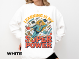 A white crewneck sweatshirt printed with a comic-style teacher superhero flying among books and school supplies under Learning Is My Super Power.