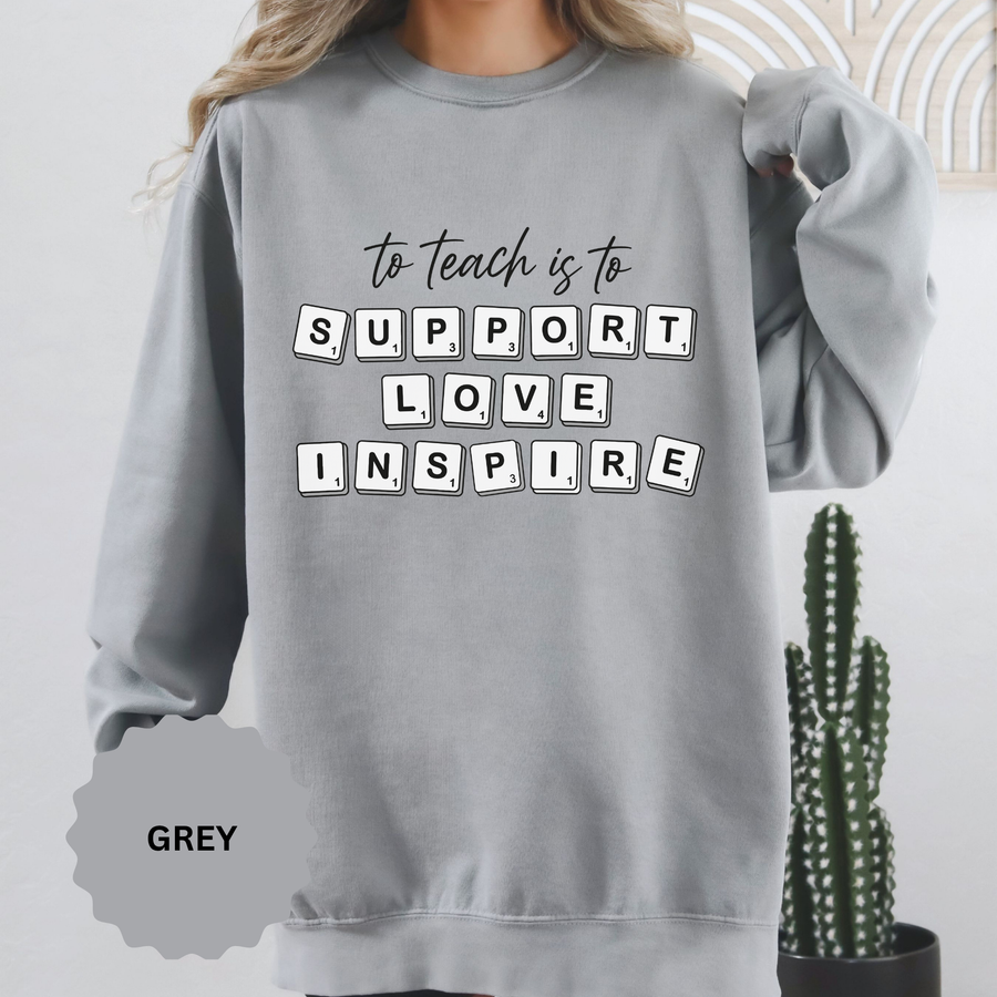 Grey sweatshirt printed with the words to teach is to in slim script above Support Love Inspire, each word spelled out in individual Scrabble-style letter tiles laid out as if on a board.