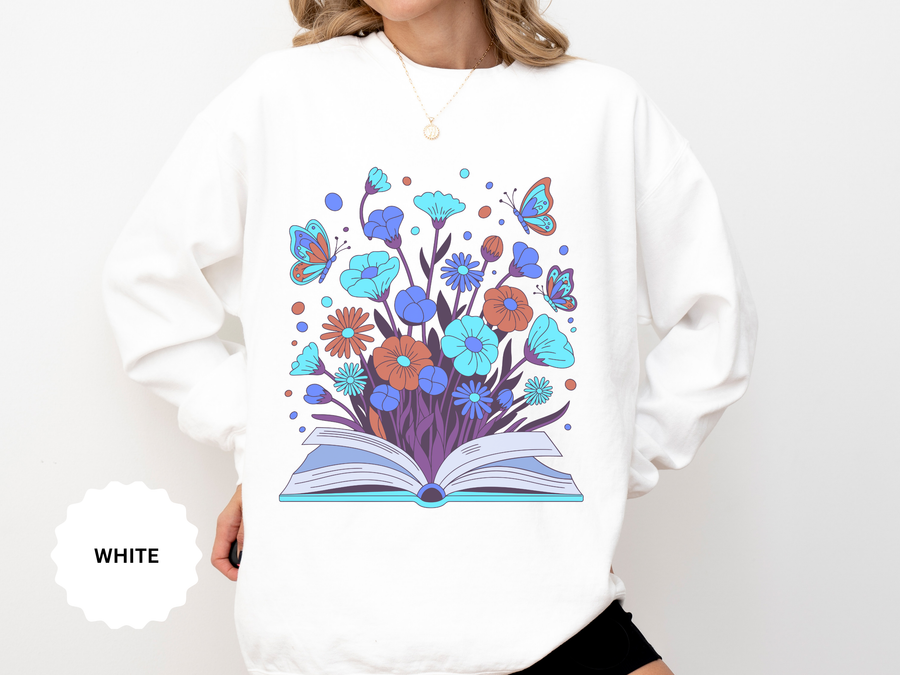 White sweatshirt printed with an open book from which blue and orange wildflowers spring upward, four butterflies rising through the blooms and a teal glow running along the open pages.