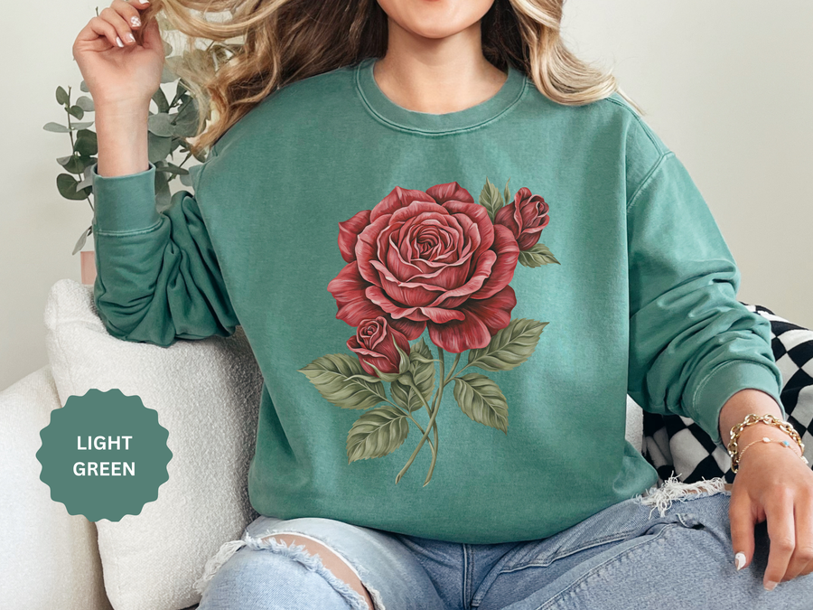 Light green sweatshirt printed with a single large red rose in botanical style, its layered petals opening above two smaller buds on a slender stem with broad sage-toned leaves.