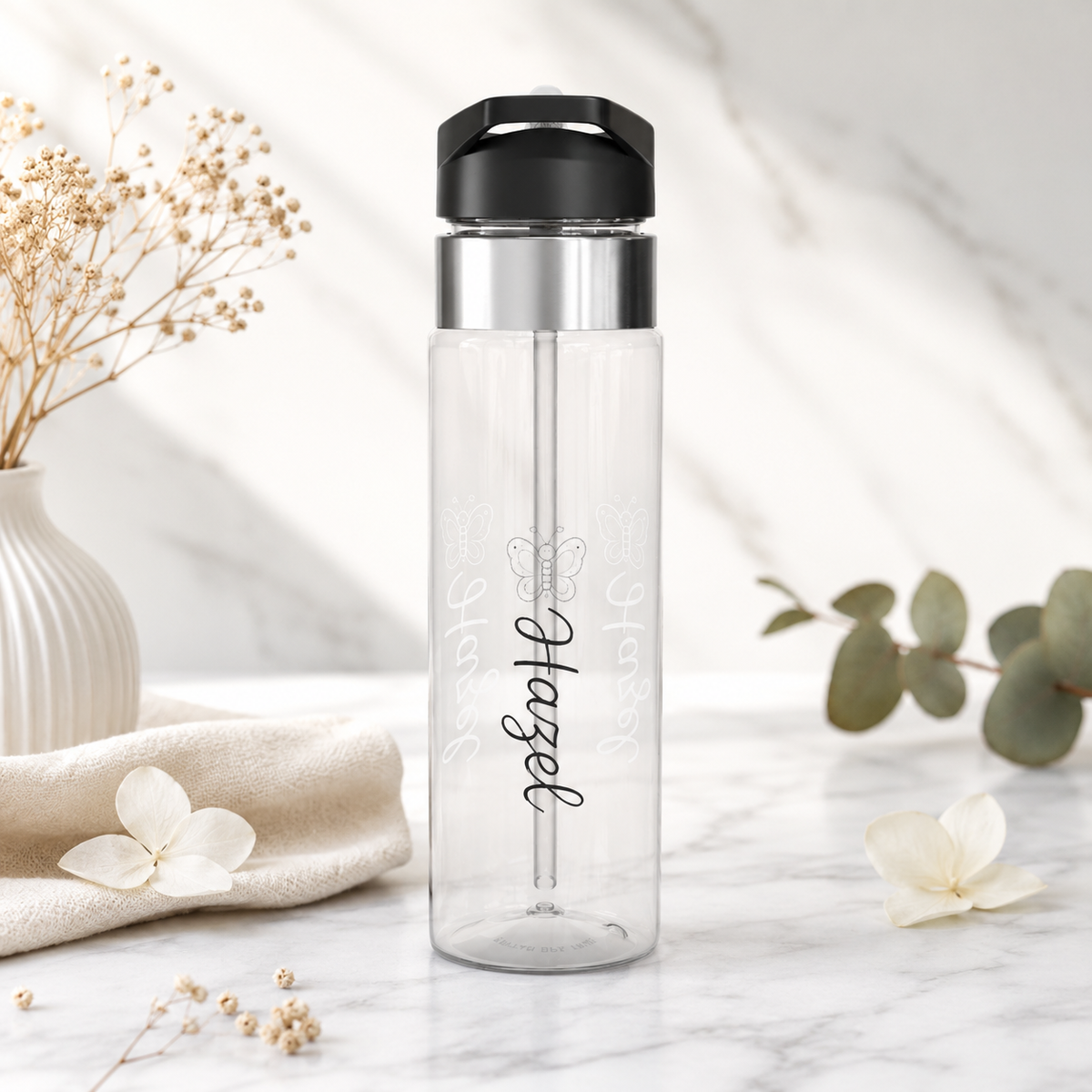 Clear plastic water bottle with a black flip lid and a straw, printed with a line-drawn butterfly above the name Hazel in black script, the name repeated faintly in white behind it.