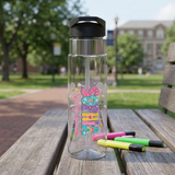 A clear plastic water bottle with a black flip-top lid sits on a wooden park bench, with colorful highlighters beside it and a blurred outdoor campus scene in the background. The bottle has a decorative multicolor graphic that reads 'Spring Break' with pastel lettering and small pastel illustrations.