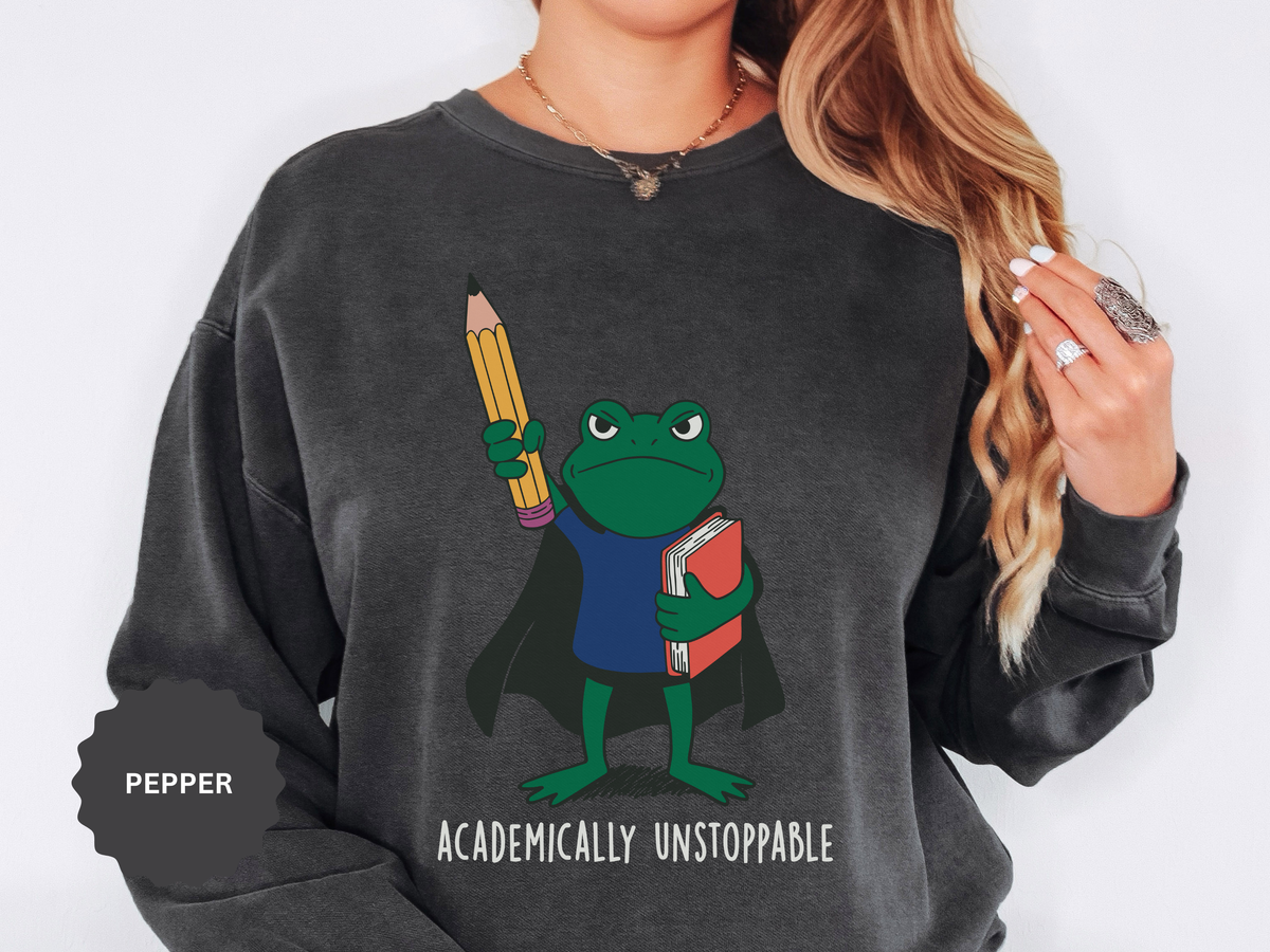 A dark gray crewneck sweatshirt printed with a frog in a cape holding a giant pencil and a book above Academically Unstoppable.