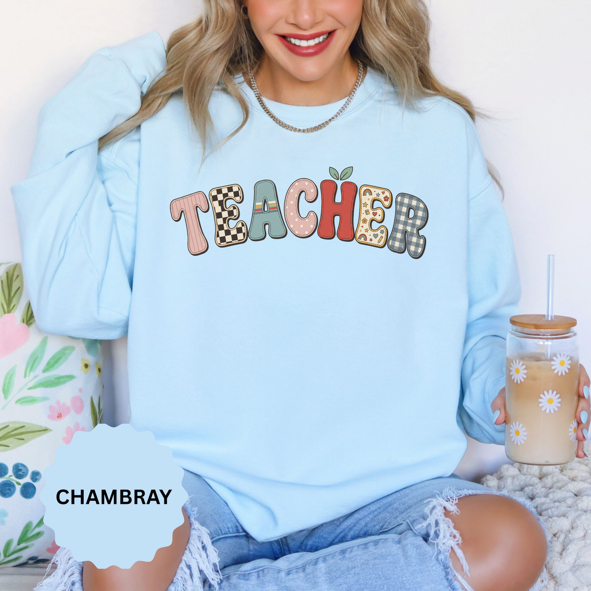 A light blue crewneck sweatshirt printed with TEACHER in patchwork letters of gingham, checkerboard and floral fabric with a small apple.