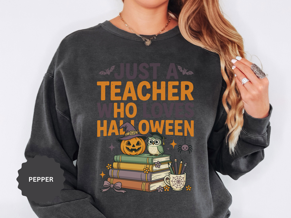 A dark gray crewneck sweatshirt printed with Just a Teacher Who Loves Halloween above a jack-o'-lantern, an owl and a stack of books.
