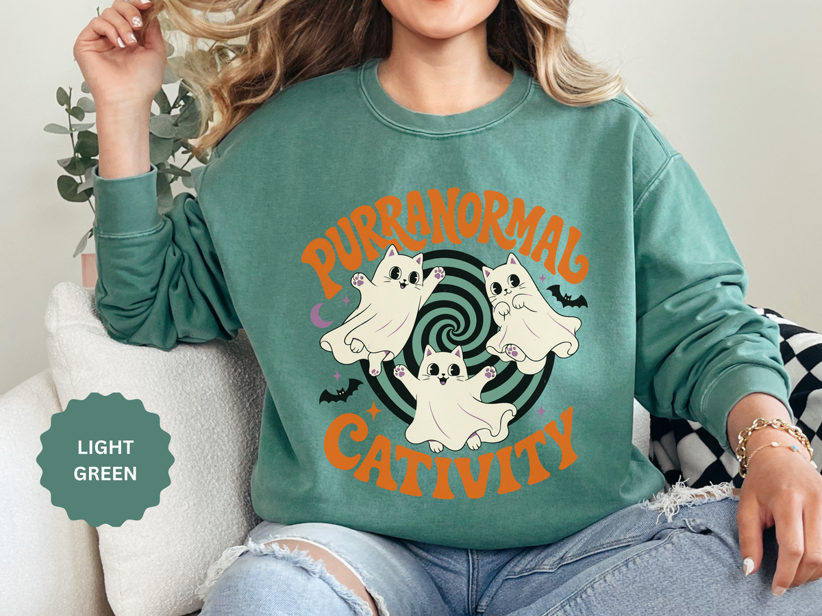 A green crewneck sweatshirt printed with three ghost cats swirling around a spiral between Purranormal and Cativity.