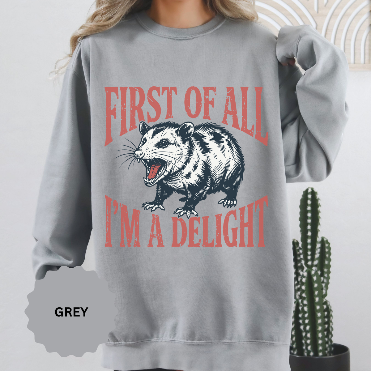 A gray crewneck sweatshirt printed with a snarling opossum between First of All and I'm a Delight in red lettering.