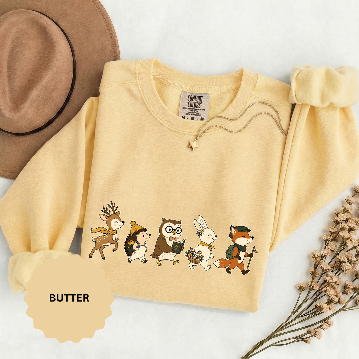 A pale yellow crewneck sweatshirt printed with a row of woodland animals, including a deer, hedgehog, owl, bunny and fox, walking with backpacks.