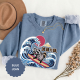 Blue jean sweatshirt folded flat with the cuffs rolled back, printed with a raccoon surfing a breaking Japanese wave below a red sun and a mountain, with Japanese characters set beside it.