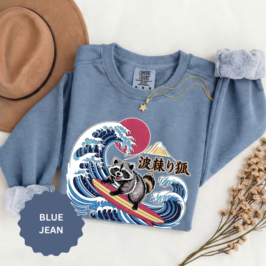 Blue jean sweatshirt folded flat with the cuffs rolled back, printed with a raccoon surfing a breaking Japanese wave below a red sun and a mountain, with Japanese characters set beside it.