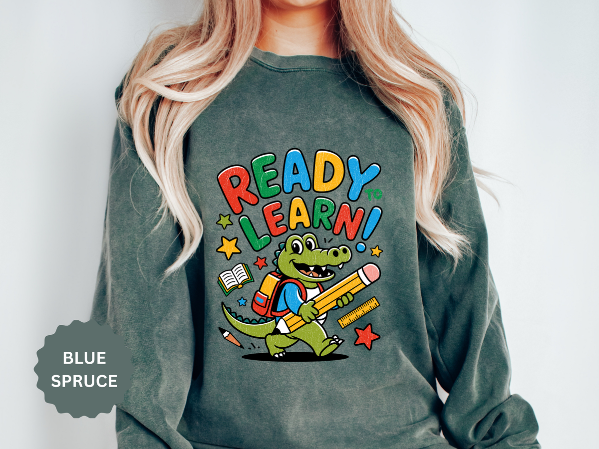 A green crewneck sweatshirt printed with a cartoon alligator in a backpack carrying a pencil under Ready to Learn! in rainbow letters.