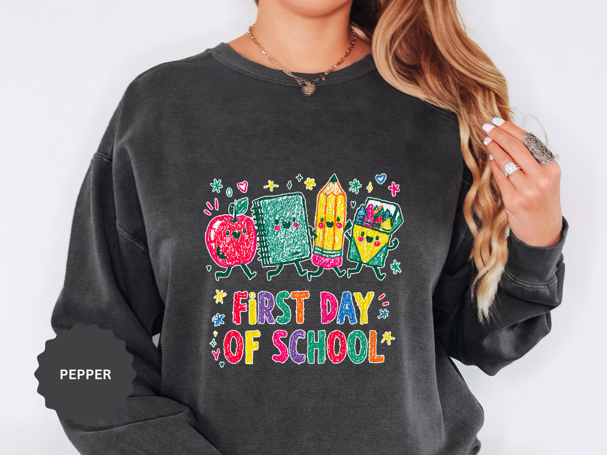 A dark gray crewneck sweatshirt printed with a cartoon apple, notebook, pencil and ruler above First Day of School in bright multicolored letters.