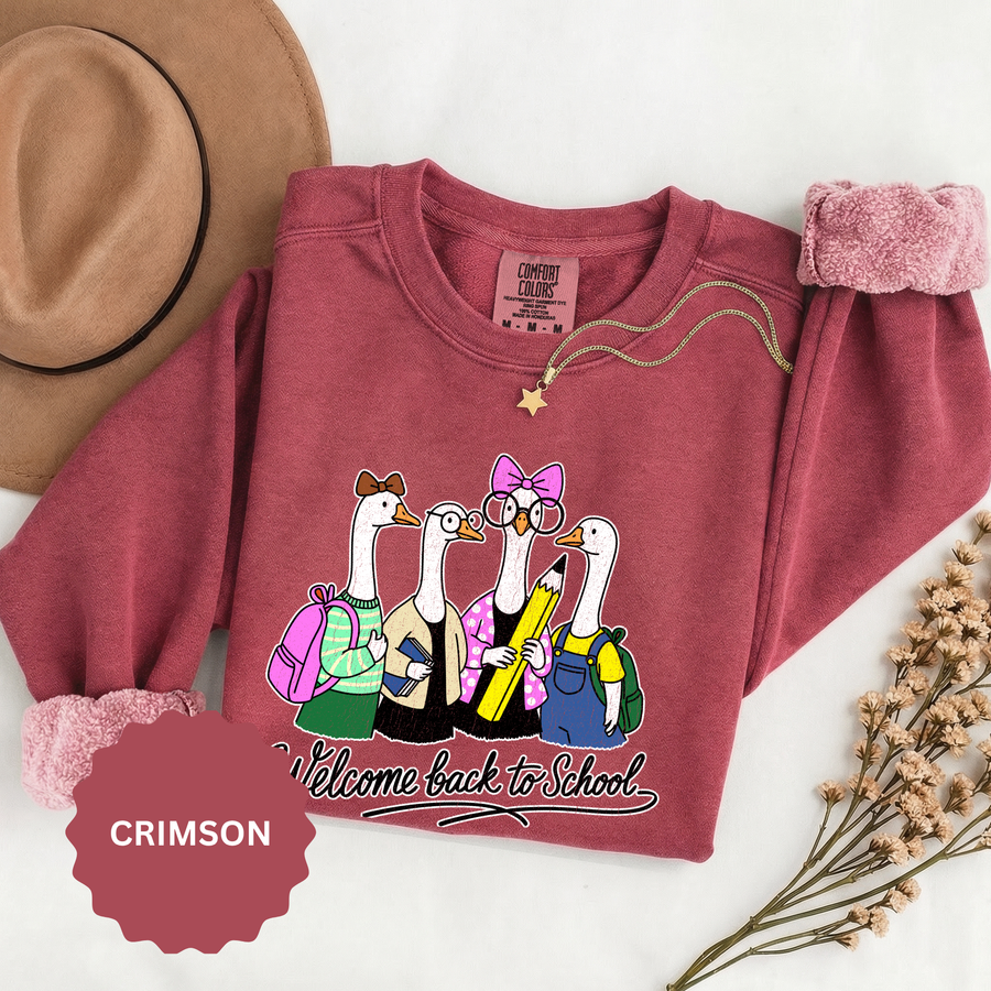 A red crewneck sweatshirt printed with four geese in backpacks, glasses and bows holding a giant pencil above Welcome back to School in script.