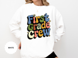 A white crewneck sweatshirt printed with First Grade Crew in retro multicolored bubble letters with small daisies.