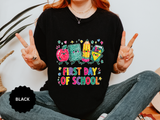 A black T-shirt printed with a cartoon apple, notebook, pencil and ruler above First Day of School in bright multicolored letters.