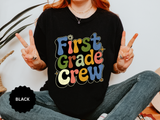 A black T-shirt printed with First Grade Crew in retro multicolored bubble letters with small daisies.
