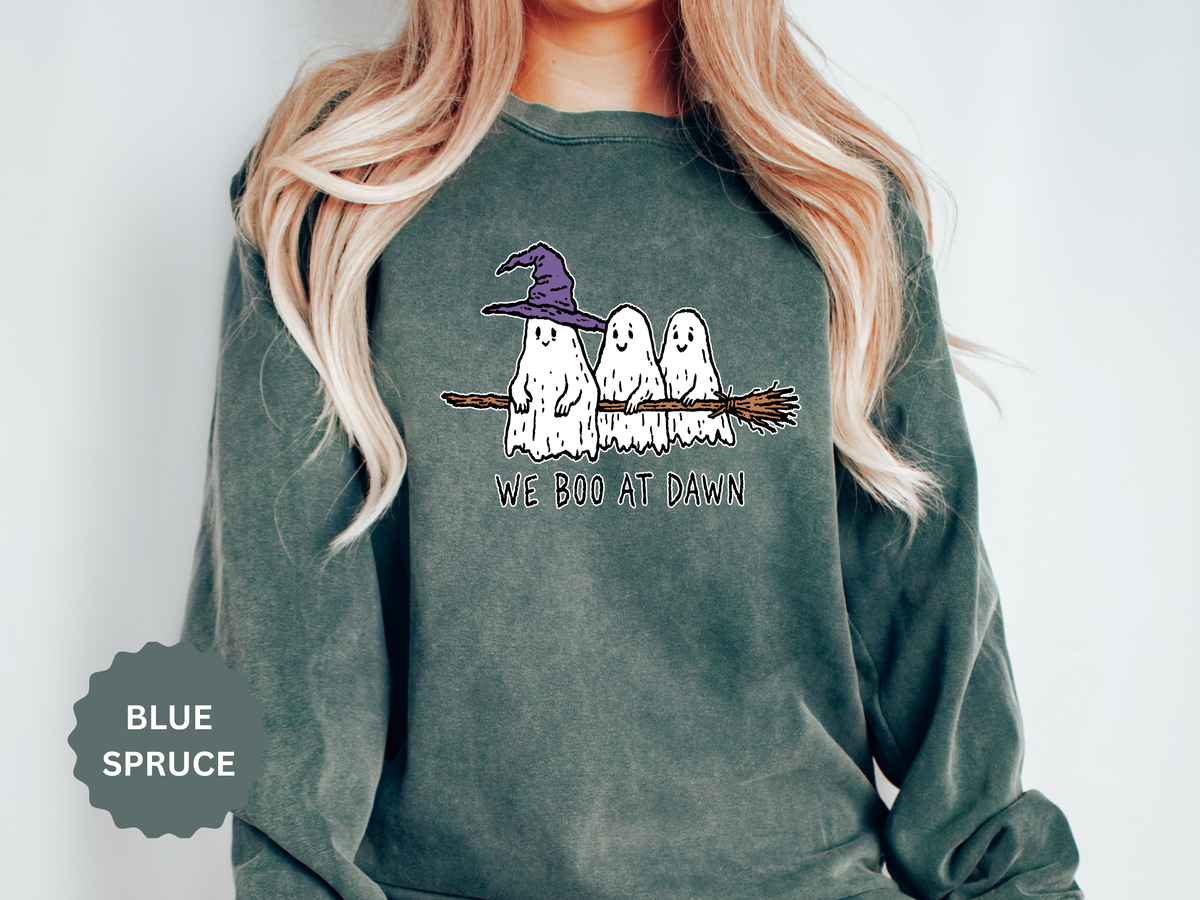 A green crewneck sweatshirt printed with three ghosts, one in a purple witch hat, sitting on a broomstick above the words We Boo at Dawn.