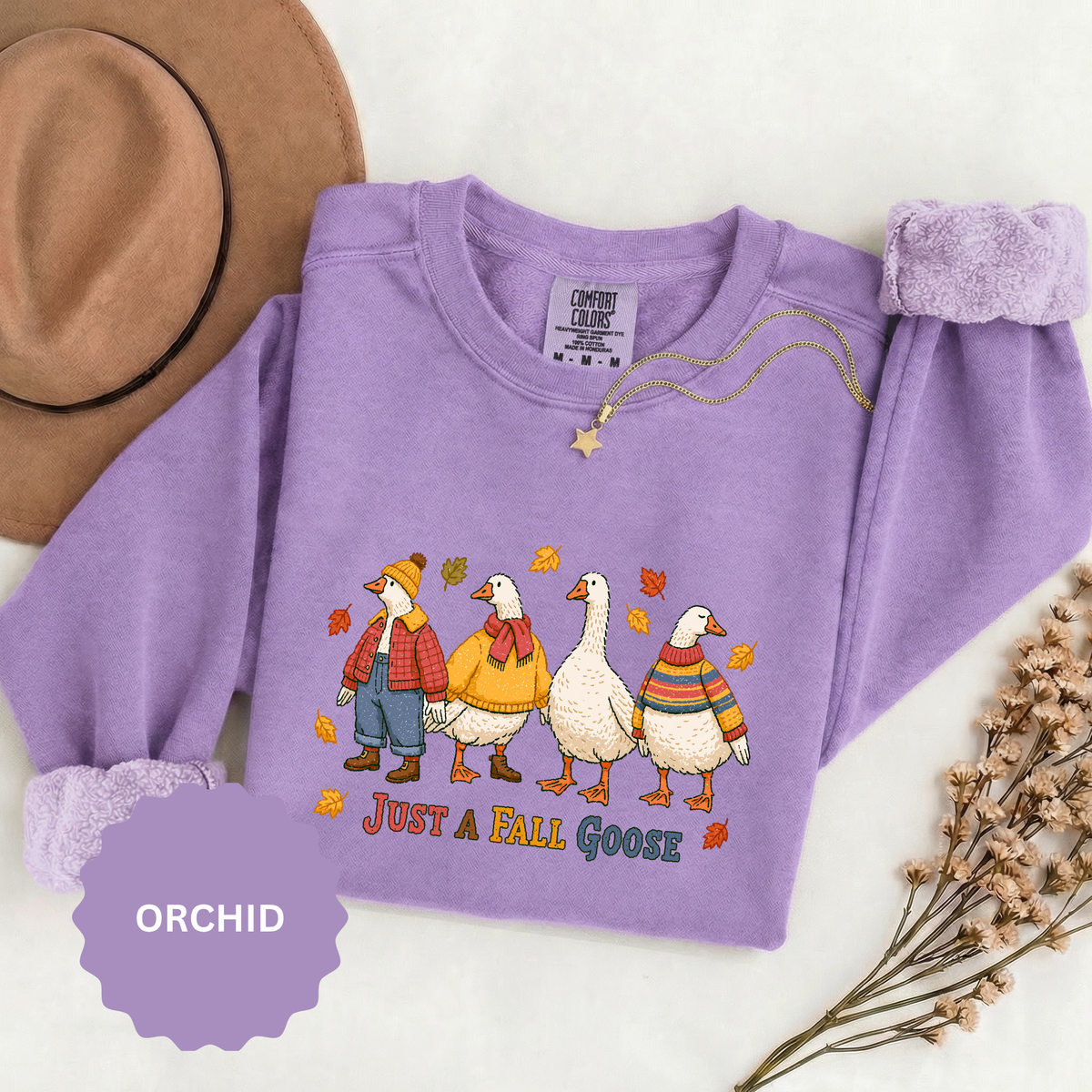 A purple crewneck sweatshirt printed with four geese dressed in autumn sweaters and scarves among falling leaves above Just a Fall Goose.