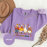 A purple crewneck sweatshirt printed with four geese dressed in autumn sweaters and scarves among falling leaves above Just a Fall Goose.