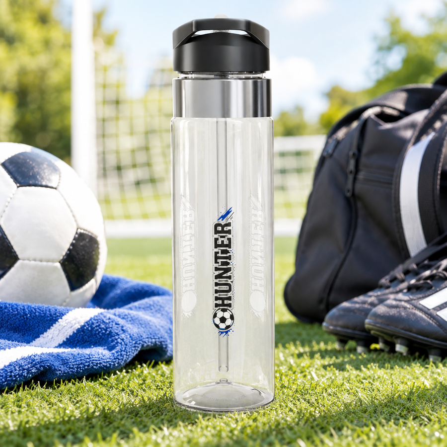 A clear Tritan water bottle with a black screw-on lid and straw, printed with soccer ball and player graphics and a name, styled with sports gear.