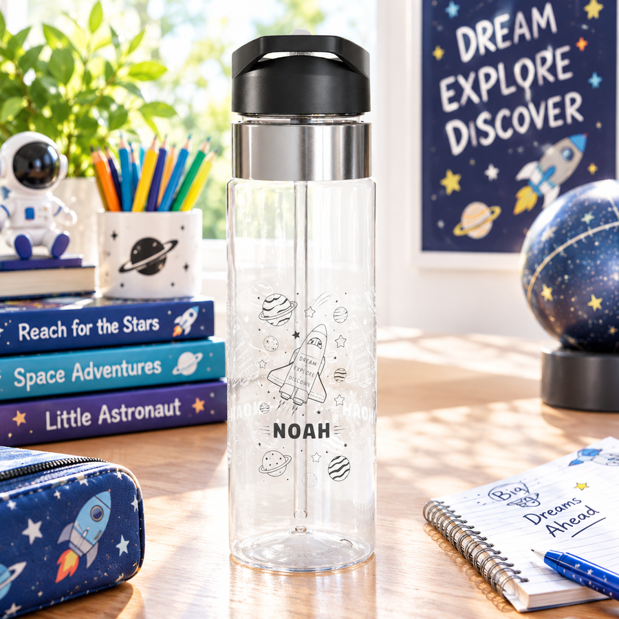 A clear Tritan water bottle with a black screw-on lid and straw, printed with rocket, planet, and star graphics and a name, styled with space-themed toys.