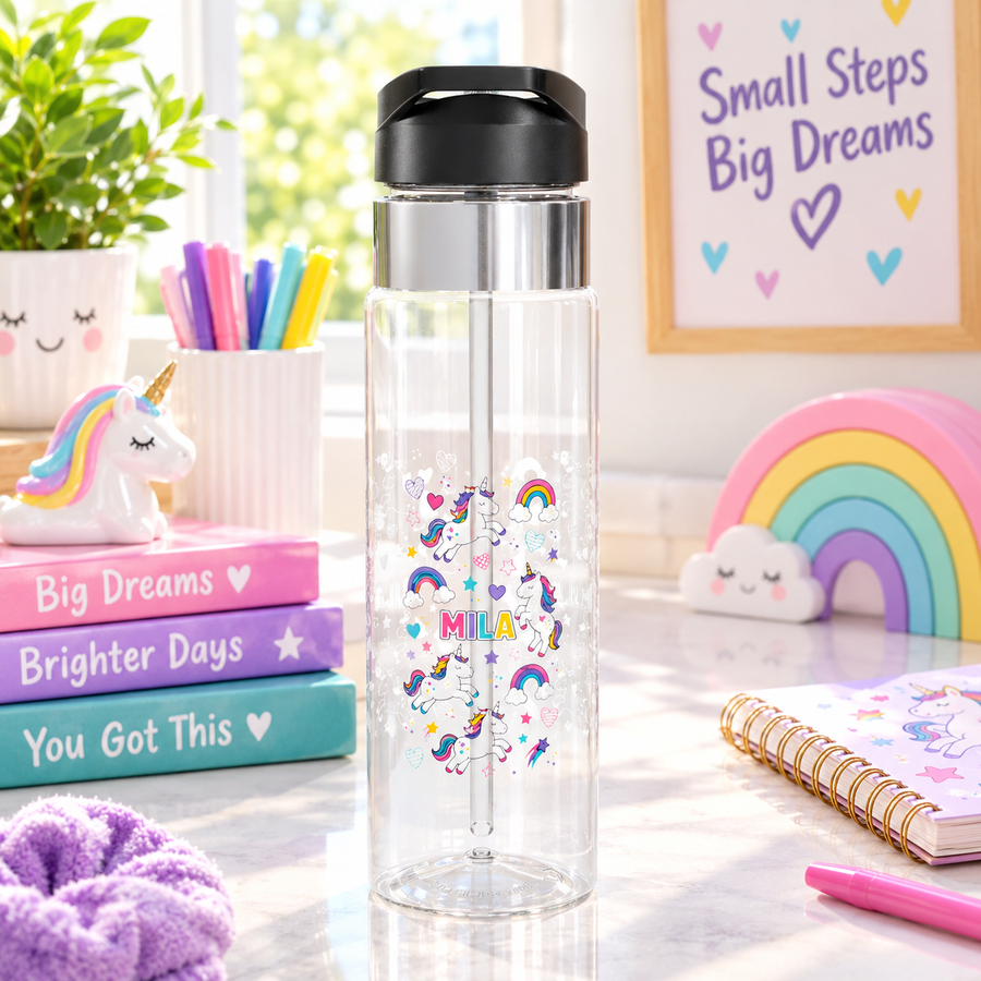 A clear Tritan water bottle with a black screw-on lid and straw, printed with colorful unicorn and rainbow graphics and a name, styled on a desk with school supplies.