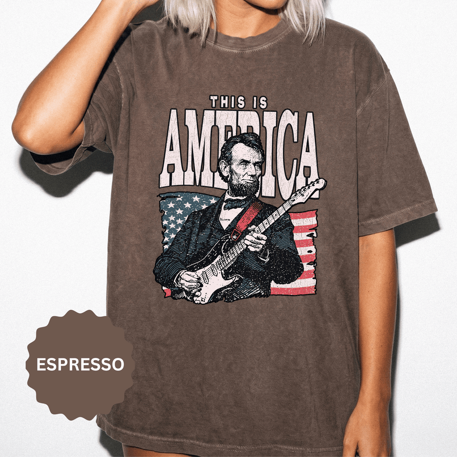 A brown T-shirt printed with an illustration of Abraham Lincoln playing an electric guitar in front of an American flag under This Is America.