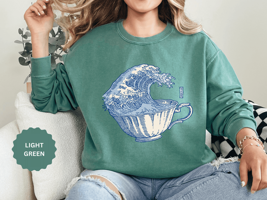 A green crewneck sweatshirt printed with a large blue and white ocean wave curling out of a teacup.