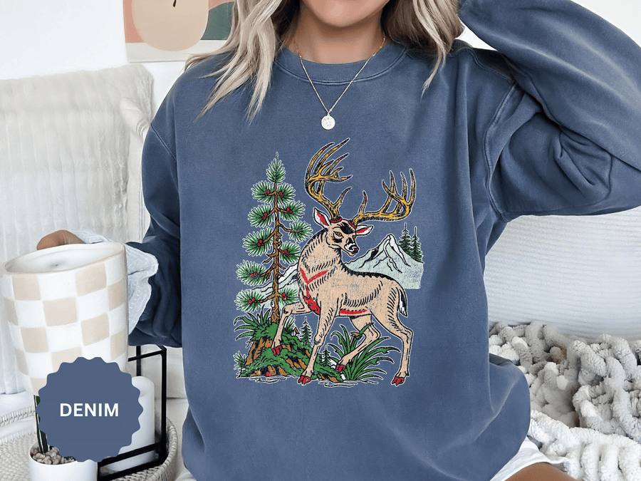 A blue crewneck sweatshirt printed with a vintage-style deer with large antlers standing beside a pine tree with mountains behind.