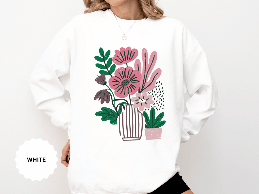 A white crewneck sweatshirt printed with pink and purple flowers in a striped vase beside a small potted plant.