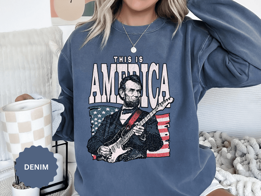 A blue crewneck sweatshirt printed with an illustration of Abraham Lincoln playing an electric guitar in front of an American flag under This Is America.