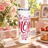 A white stainless steel skinny tumbler with a clear straw lid, printed with a pink heart-shaped word design reading "MOM" and phrases like "my greatest love", on a table.
