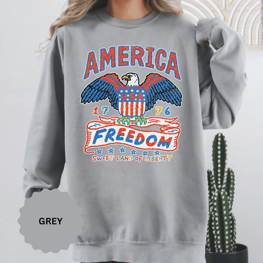An orange crewneck sweatshirt printed with a bald eagle and flag shield between America, 1776, Freedom and Sweet Land of Liberty.
